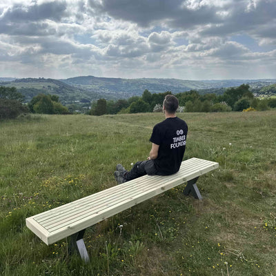 Man sitting on a wooden garden bench with steel x legs with rolling hills in the background, part of the Wooden Garden Dining Set.