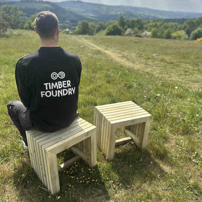 Man sitting on a wooden garden stool in the sunshine with rolling hills in the background, part of the Wooden Garden Dining Set.