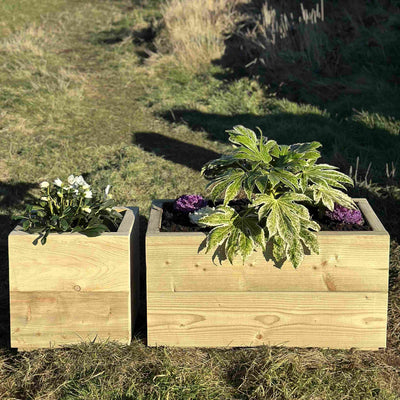 Heavy-duty wooden trough planters, one square and one rectangle filled with green plants and purple and white flowers. Placed on grass in front of a stone wall.