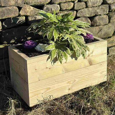 Heavy-duty wooden trough planter filled with a green plant and purple and white flowers. Placed on grass in front of a stone wall.