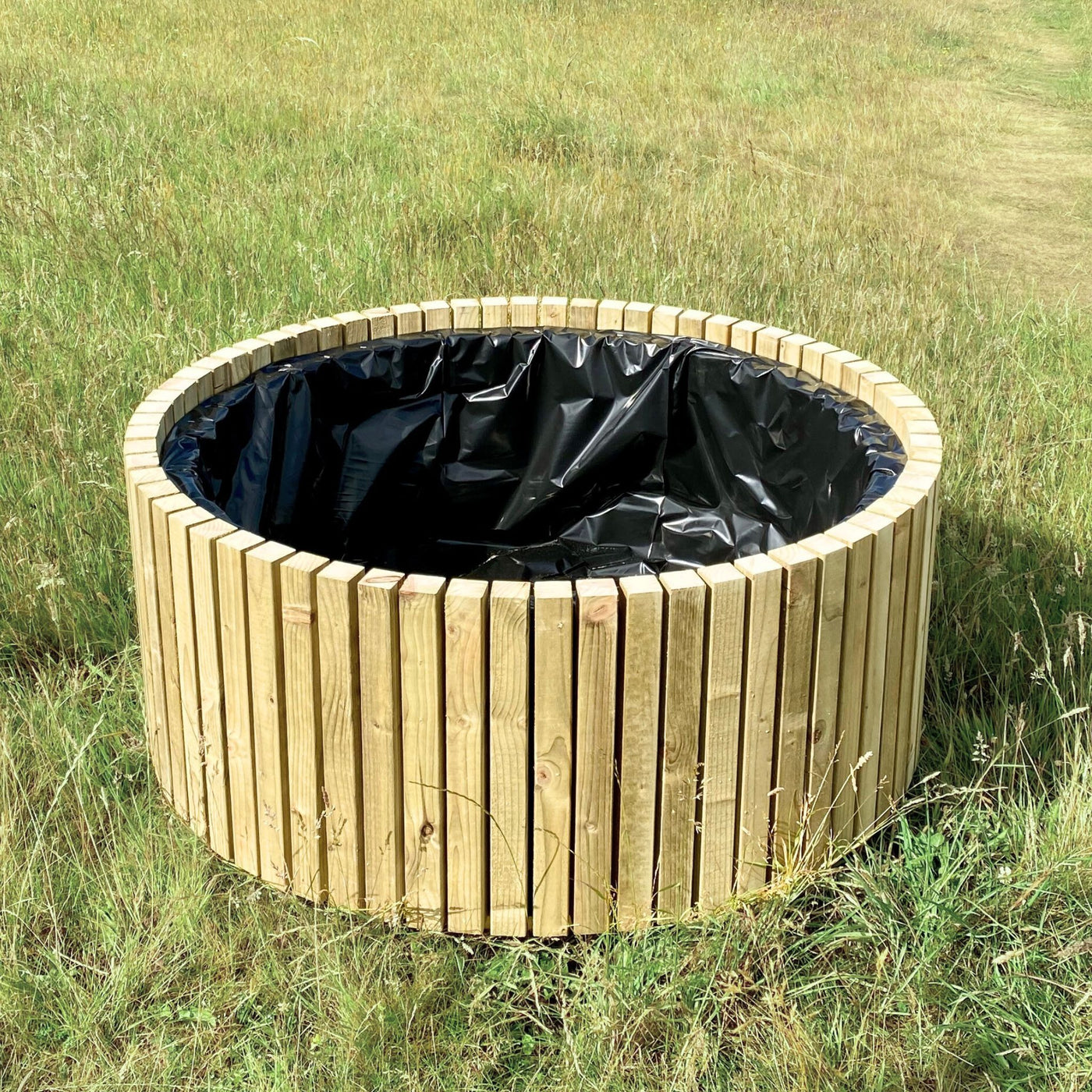 Wooden circular planter with black liner in a grassy field. Large round wood garden planter with lining, made from sustainable timber in the UK.
