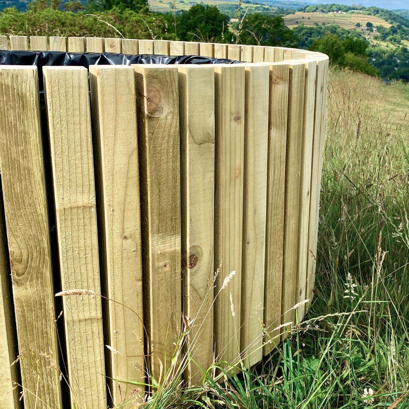 Wooden planter in a natural setting with grass and trees. The large wooden barrel-shaped planter is delivered fully assembled and lined.