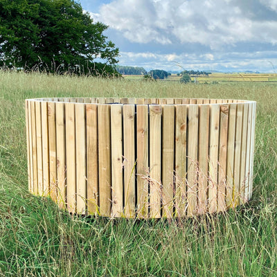 Wooden planter in a grassy field with a scenic background. The barrel style wooden planter is made form sustainable timber and crafted in the UK.