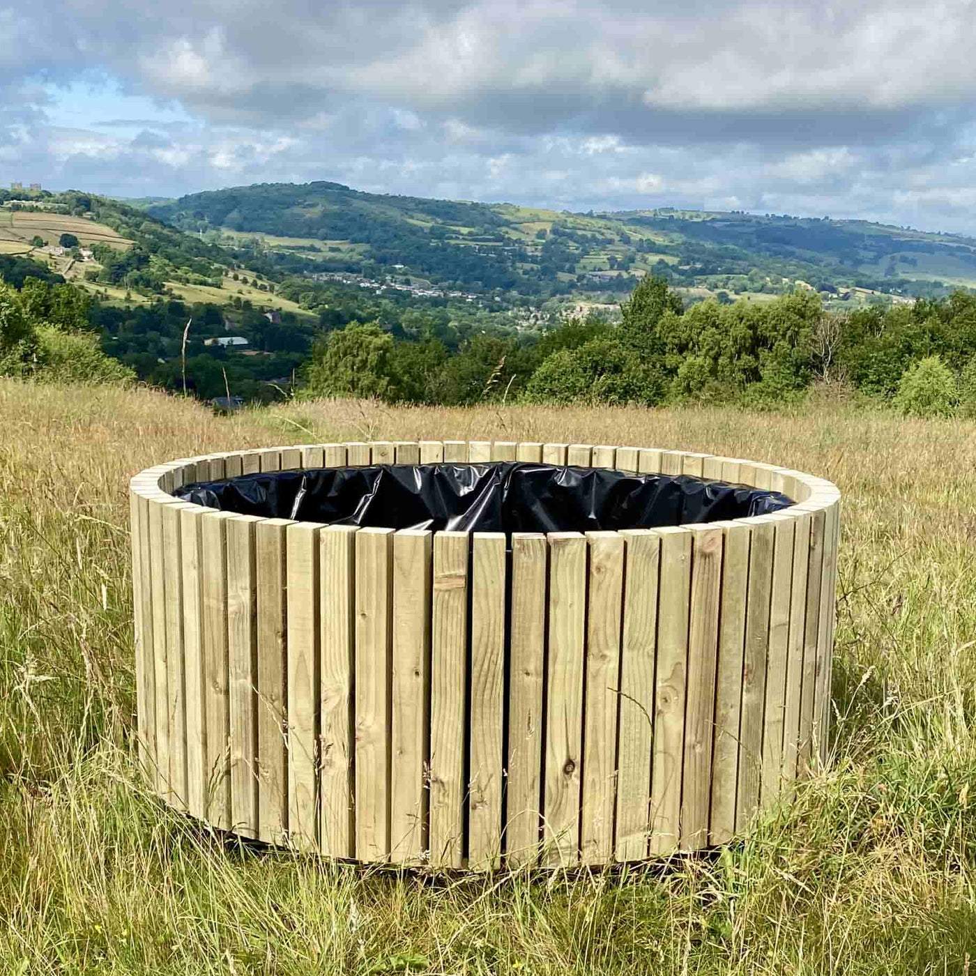 Wooden round planter in a grassy field with a scenic view of hills and sky. The large wooden planter is delivered fully assembled and lined.